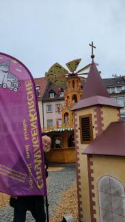 Rechts die Wagenkirche mit Turm, links eine Beachflag mit Aufschrift "CitykWagenKirche - schnell mal Kirche". In Hintergrund die große Weinachtspyramide des Weihnachtsmarkts. Lorenz Hummel lugt lachend hinter der Fahne vor.